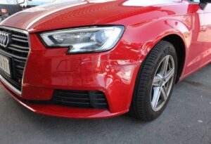 Das Bild zeigt einen roten Audi mit einem deutlich sichtbaren Schaden an der linken vorderen Karosserie. Die Lackierung ist zerkratzt und eingedellt, was auf einen kürzlichen Unfall hindeutet.